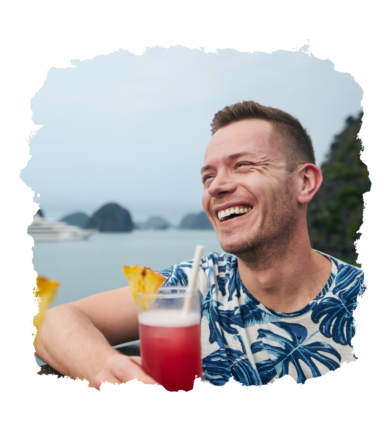Lächelnder Mann mit einem Cocktail in der Hand, im Hintergrund eine Küstenlandschaft.