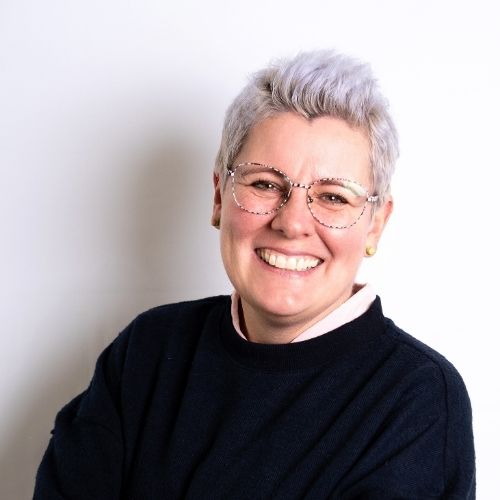 Lächelnde Person mit grauen Haaren, Brille und schwarzem Pullover vor hellem Hintergrund.