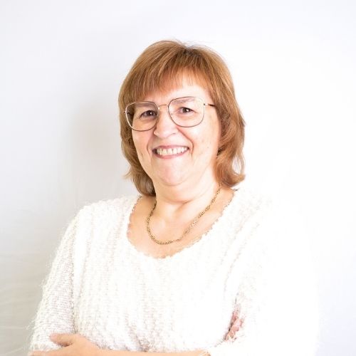 Eine Frau mit Brille und blonden Haaren, die in einer hellen Bluse lächelt und die Arme verschränkt.