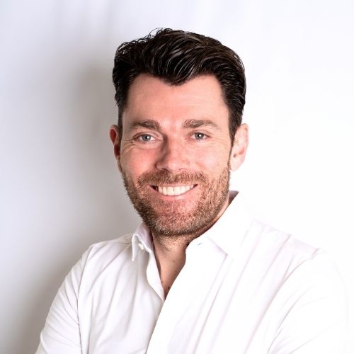 Mann mit Bart und lächelndem Gesicht in einem weißen Hemd vor einem neutralen Hintergrund.