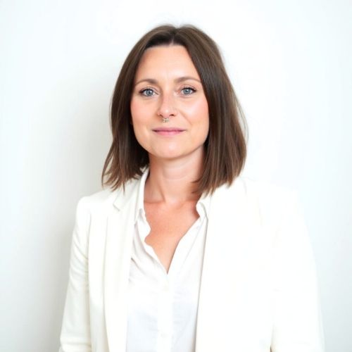 Frau mit kurzen, braunen Haaren und einem weißen Blazer vor einem neutralen Hintergrund.