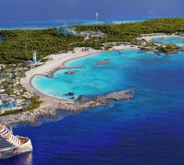 Ein Kreuzfahrtschiff liegt im Wasser vor einer Karibikinsel mit klarem, blauem Wasser und Sandstrand.