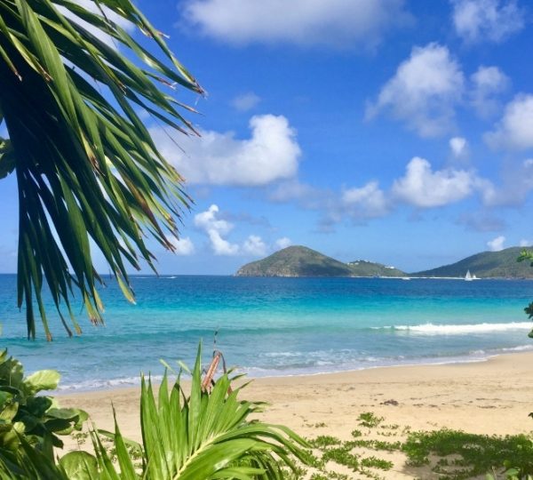 Strand mit klarem Wasser, sanften Wellen und grünen Hügeln im Hintergrund.